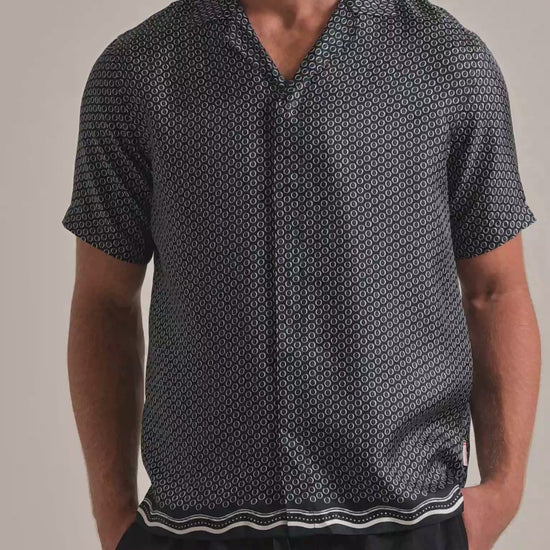 Man wearing navy printed shirt with geometric print and wavey border. Worn with dark trousers against a plain background