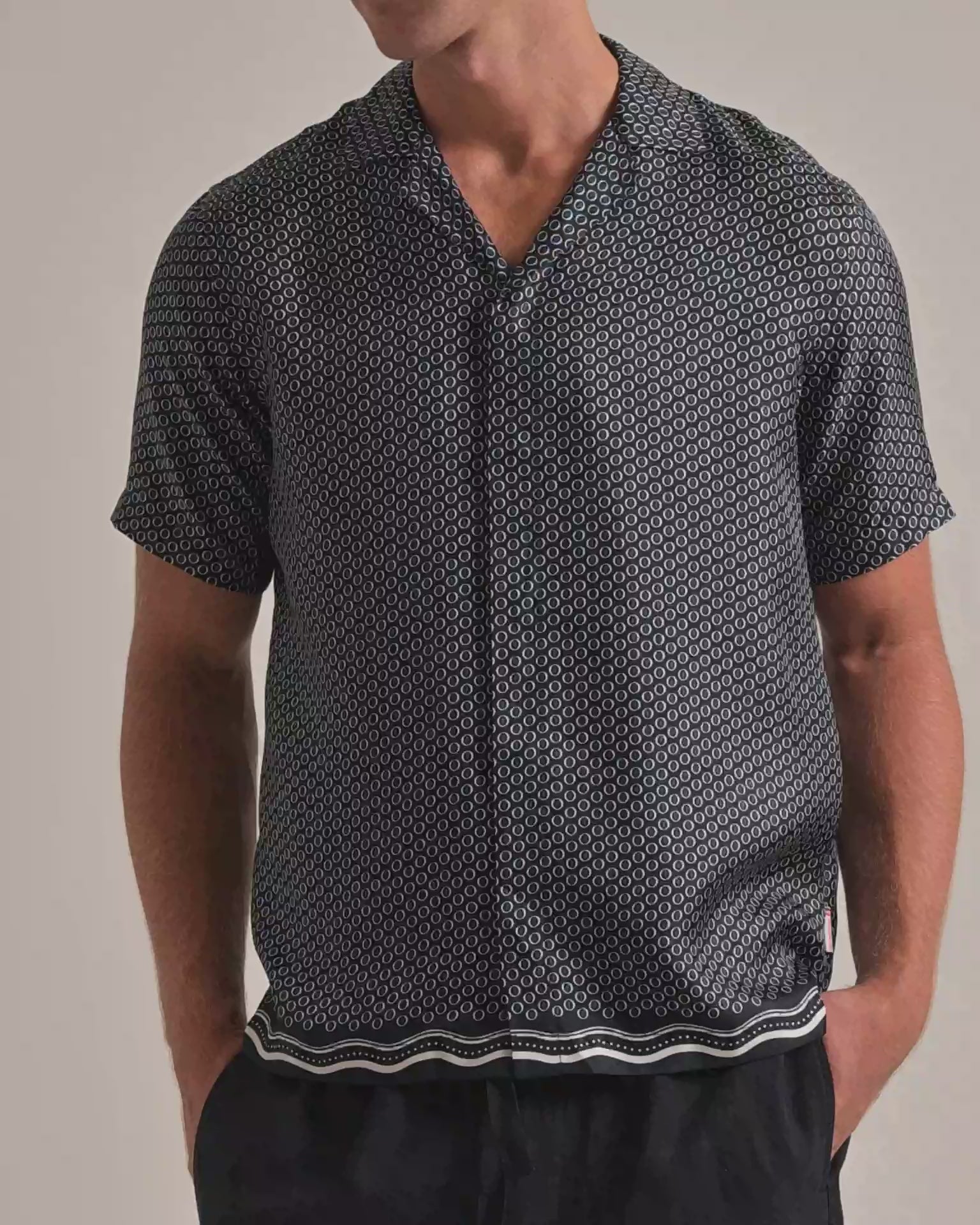 Man wearing navy printed shirt with geometric print and wavey border. Worn with dark trousers against a plain background