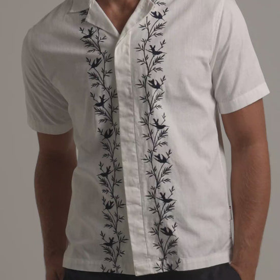 Man wearing white linen shirt with black botanical border embroidery with dark trousers against a plain background