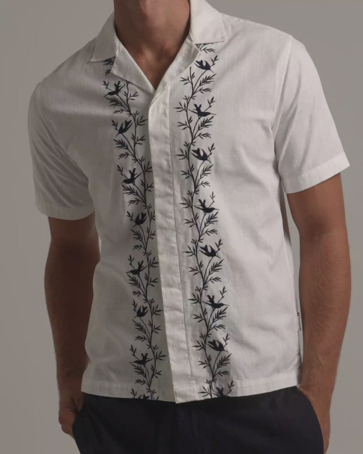 Man wearing white linen shirt with black botanical border embroidery with dark trousers against a plain background