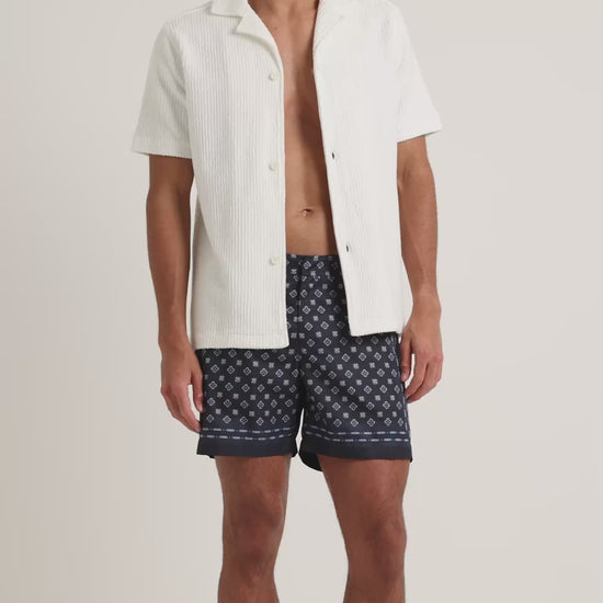 Man wearing textured white shirt with navy geometric print shorts on a plain background