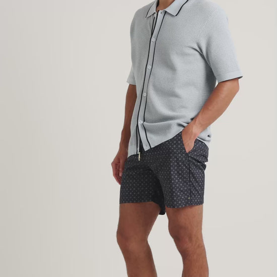 Man wearing textured knitted shirt in light blue with black trim, with geometric print navy swim shorts on a plain background
