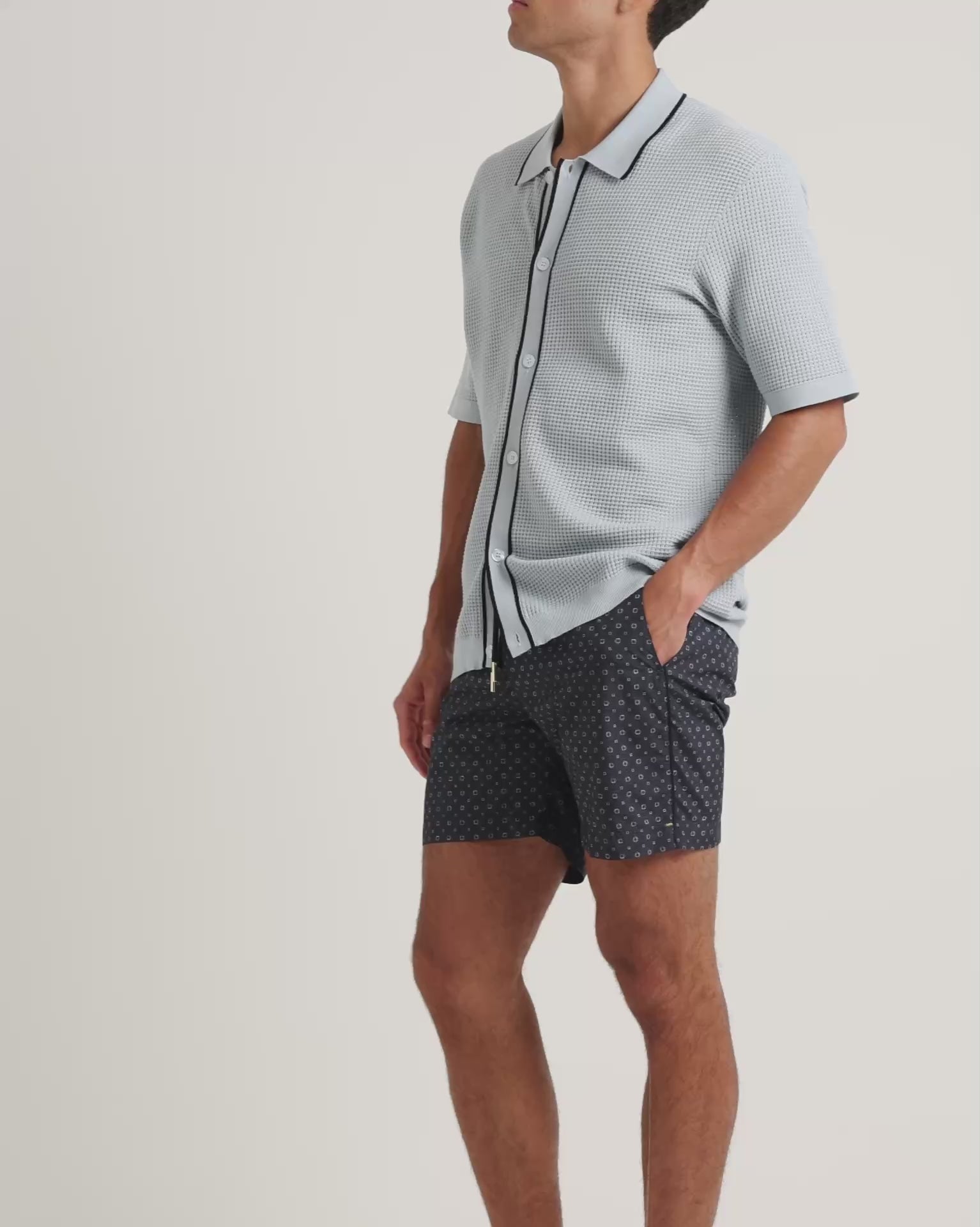 Man wearing textured knitted shirt in light blue with black trim, with geometric print navy swim shorts on a plain background