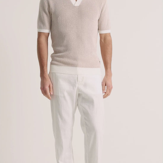 Man wearing beige crochet polo shirt with white linen trousers on a plain backgroud