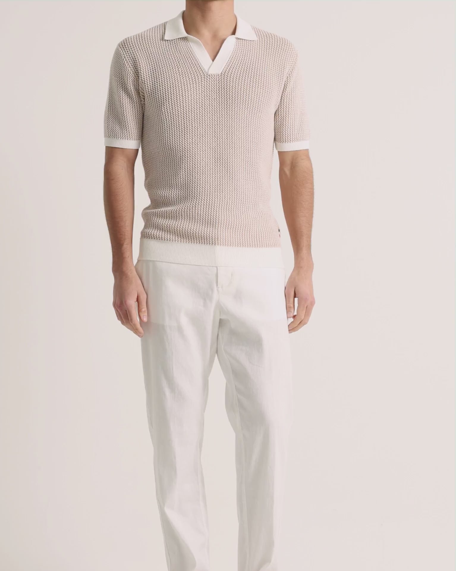 Man wearing beige crochet polo shirt with white linen trousers on a plain backgroud