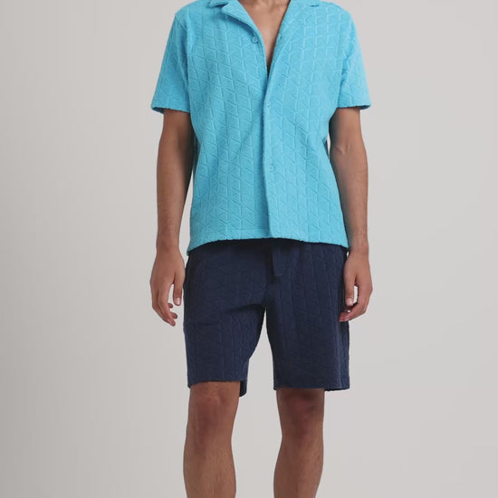 Man wearing blue towelling shirt with navy towelling shorts on a plain background