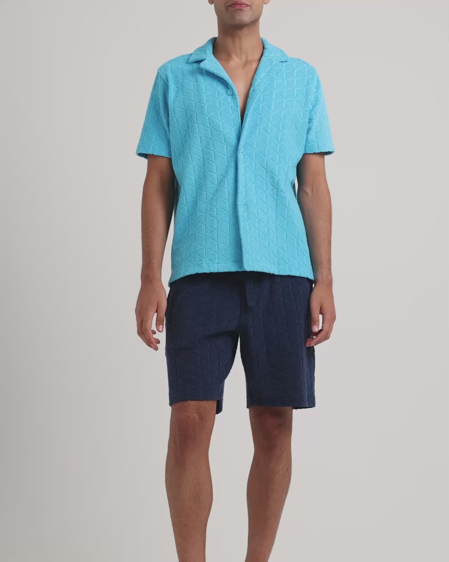 Man wearing blue towelling shirt with navy towelling shorts on a plain background