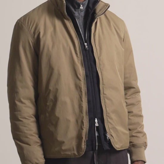 Man wearing dark beige coloured jacket layered with a black zip up and grey quarter zip. Worn with dark brown trousers against a plain background
