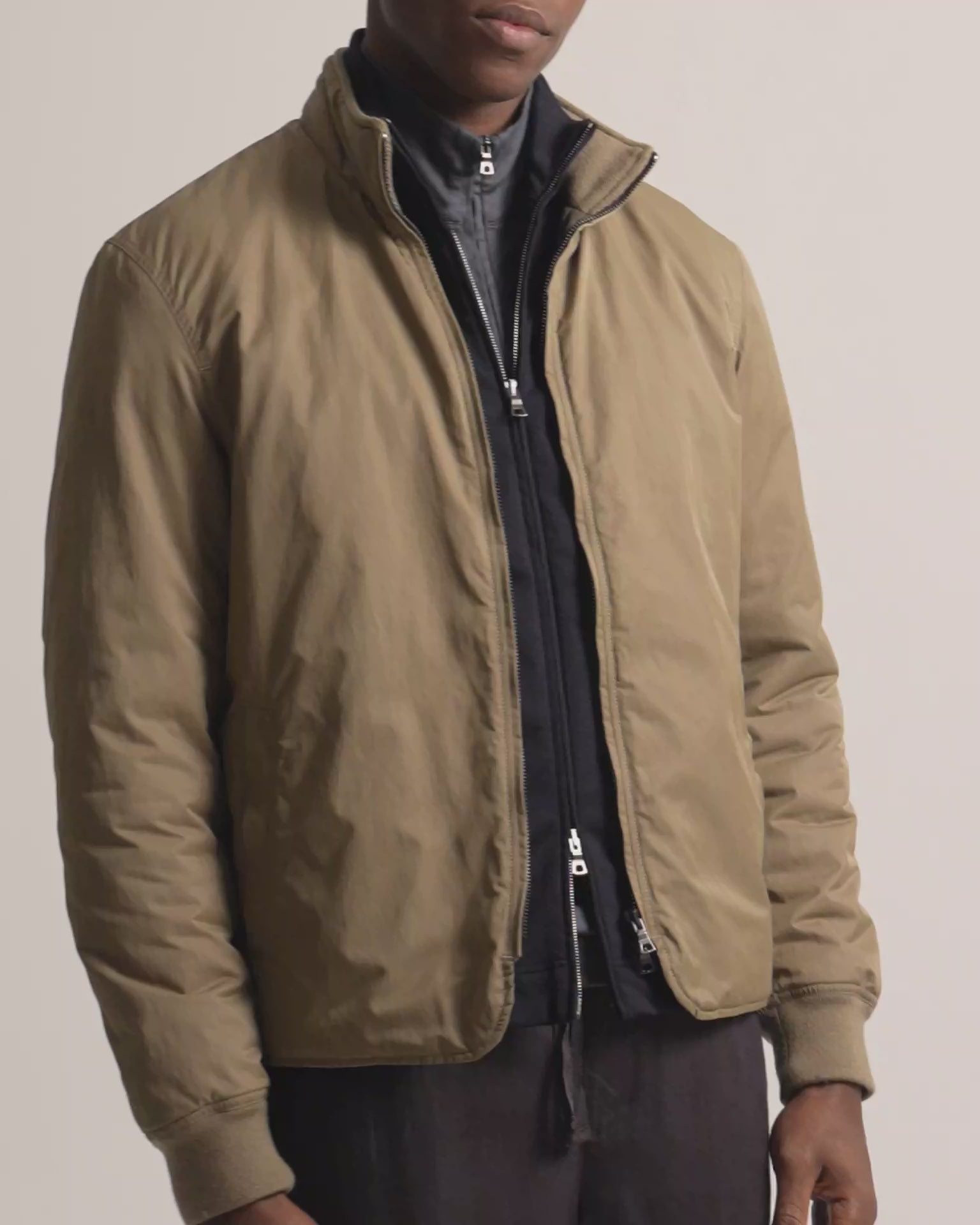 Man wearing dark beige coloured jacket layered with a black zip up and grey quarter zip. Worn with dark brown trousers against a plain background