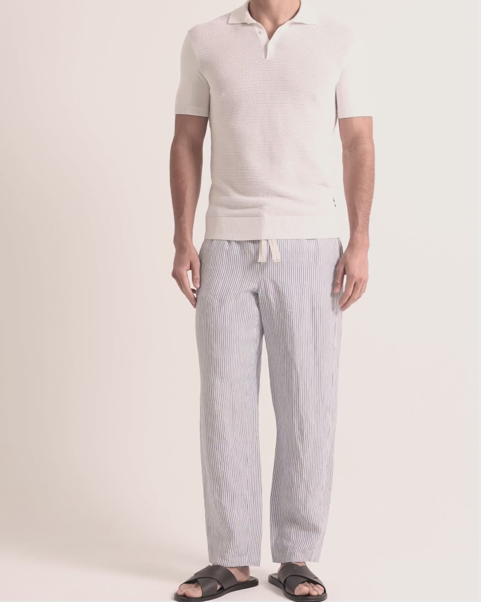 Man wearing white knitted polo shirt with blue striped trousers on a plain background