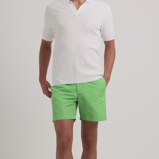 Man wearing white polo shirt with green swim shorts on a plain background