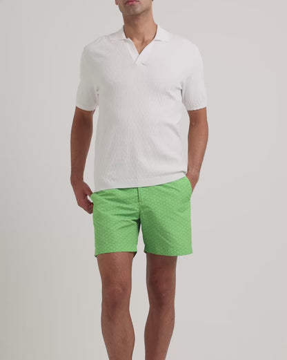 Man wearing white polo shirt with green swim shorts on a plain background