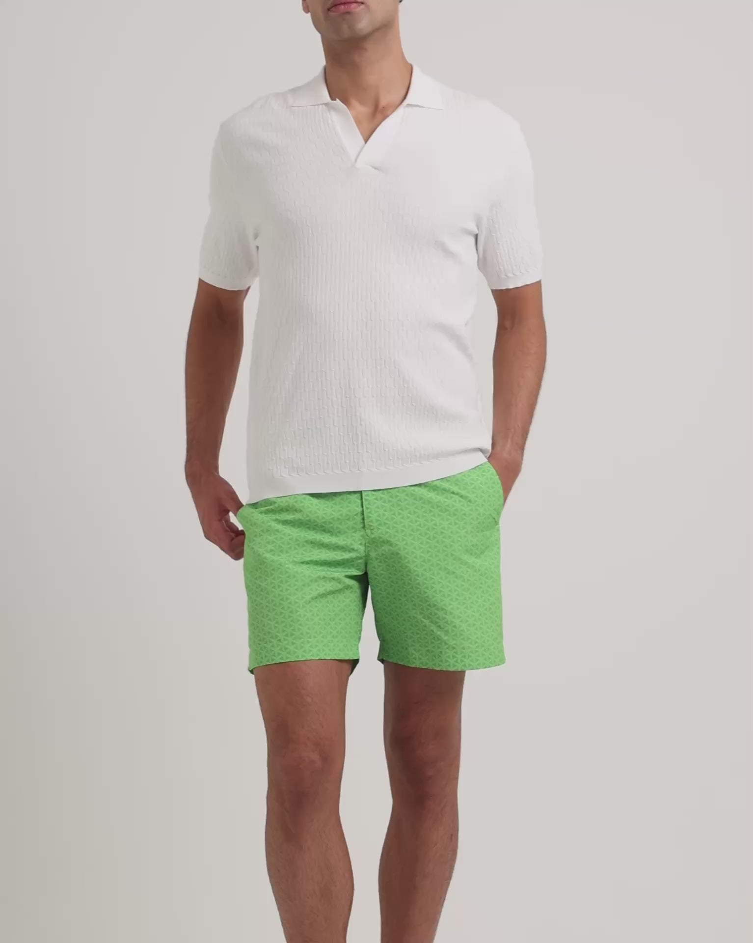 Man wearing white polo shirt with green swim shorts on a plain background