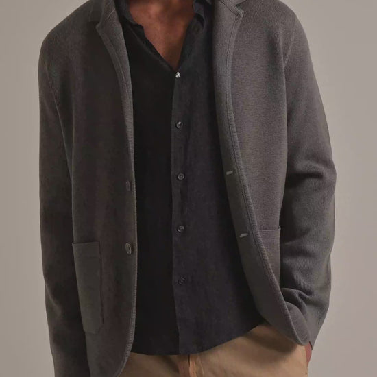 Man wearing dark grey knitted blazer with grey linen shirt and beige coloured trousers, modelling against a plain background