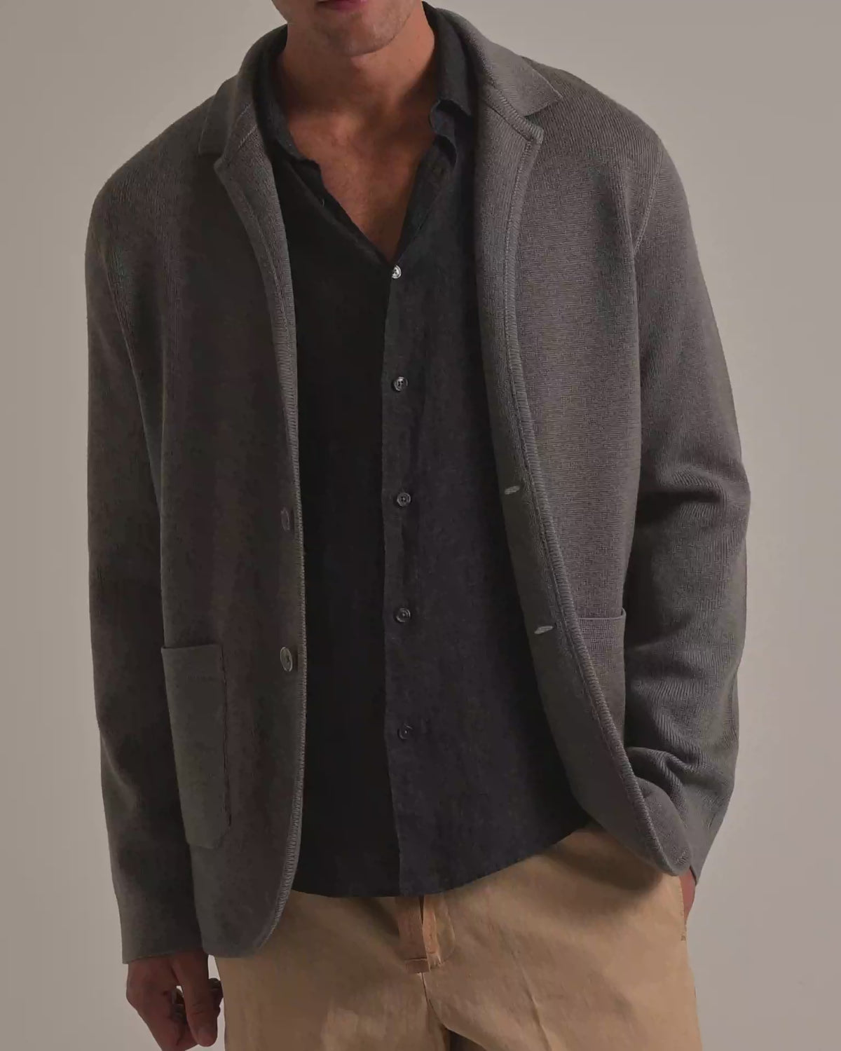 Man wearing dark grey knitted blazer with grey linen shirt and beige coloured trousers, modelling against a plain background