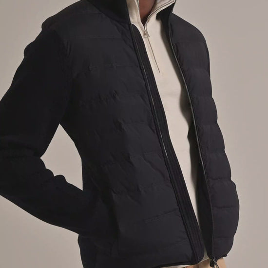 Man wearing navy padded jacket with knitted sleeves with cream quarter zip underneath and beige trousers, on a plain background