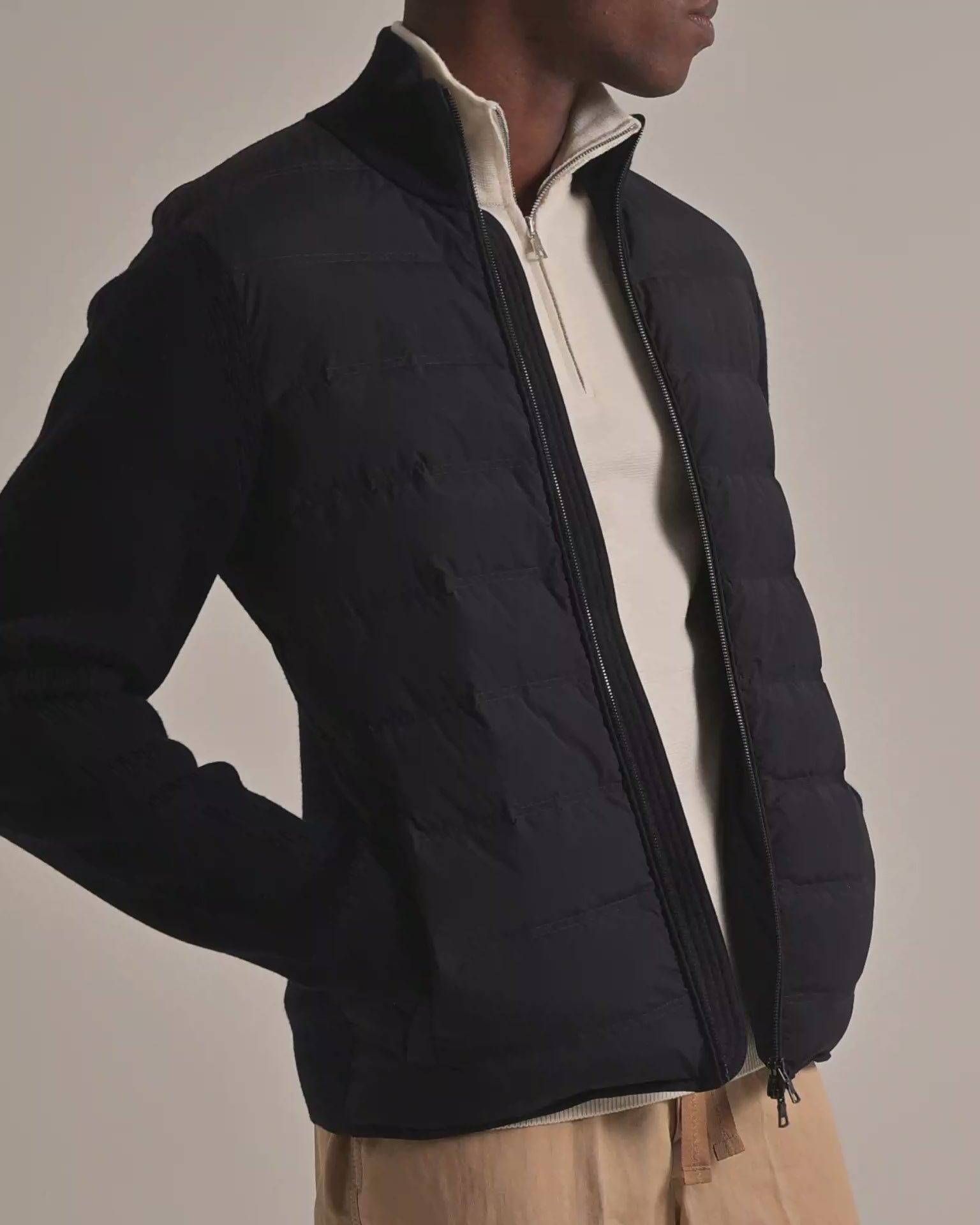 Man wearing navy padded jacket with knitted sleeves with cream quarter zip underneath and beige trousers, on a plain background