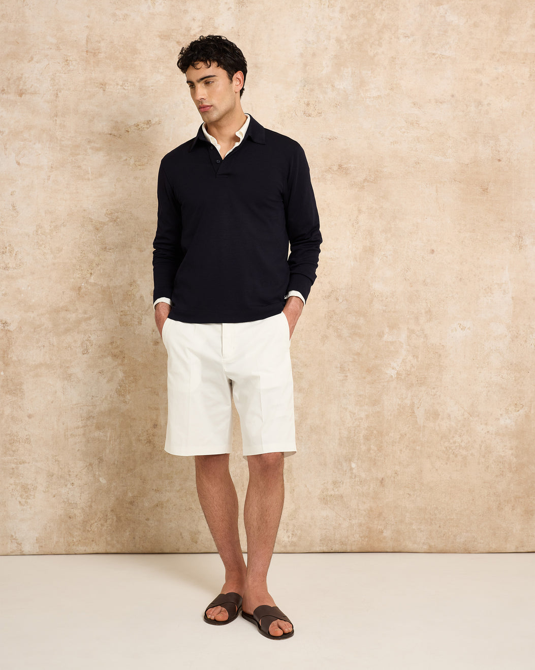 Man wearing a long sleeve polo shirt and white shorts against a beige wall