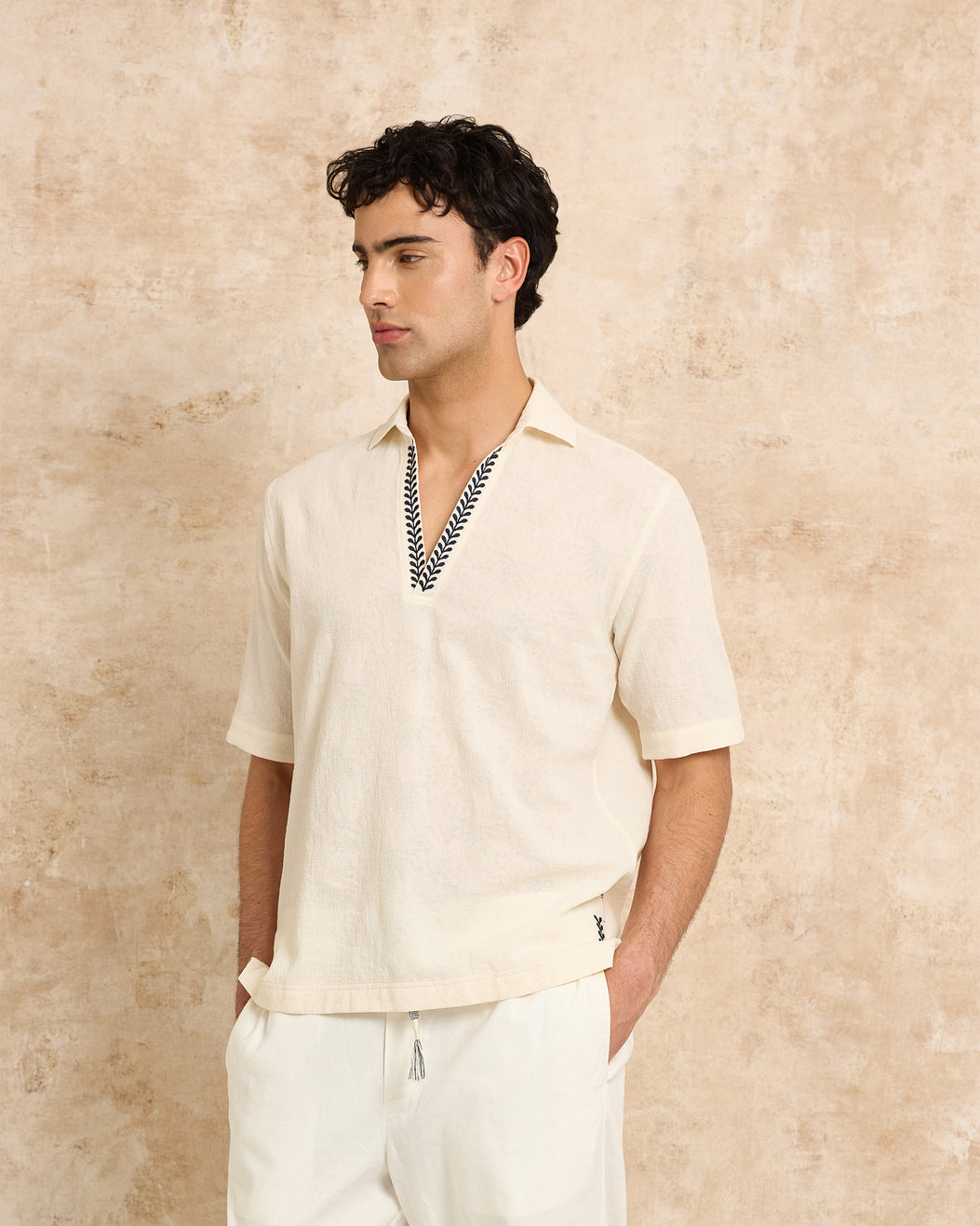 Man wearing a beige short-sleeve shirt with embroidered details against a textured beige wall.