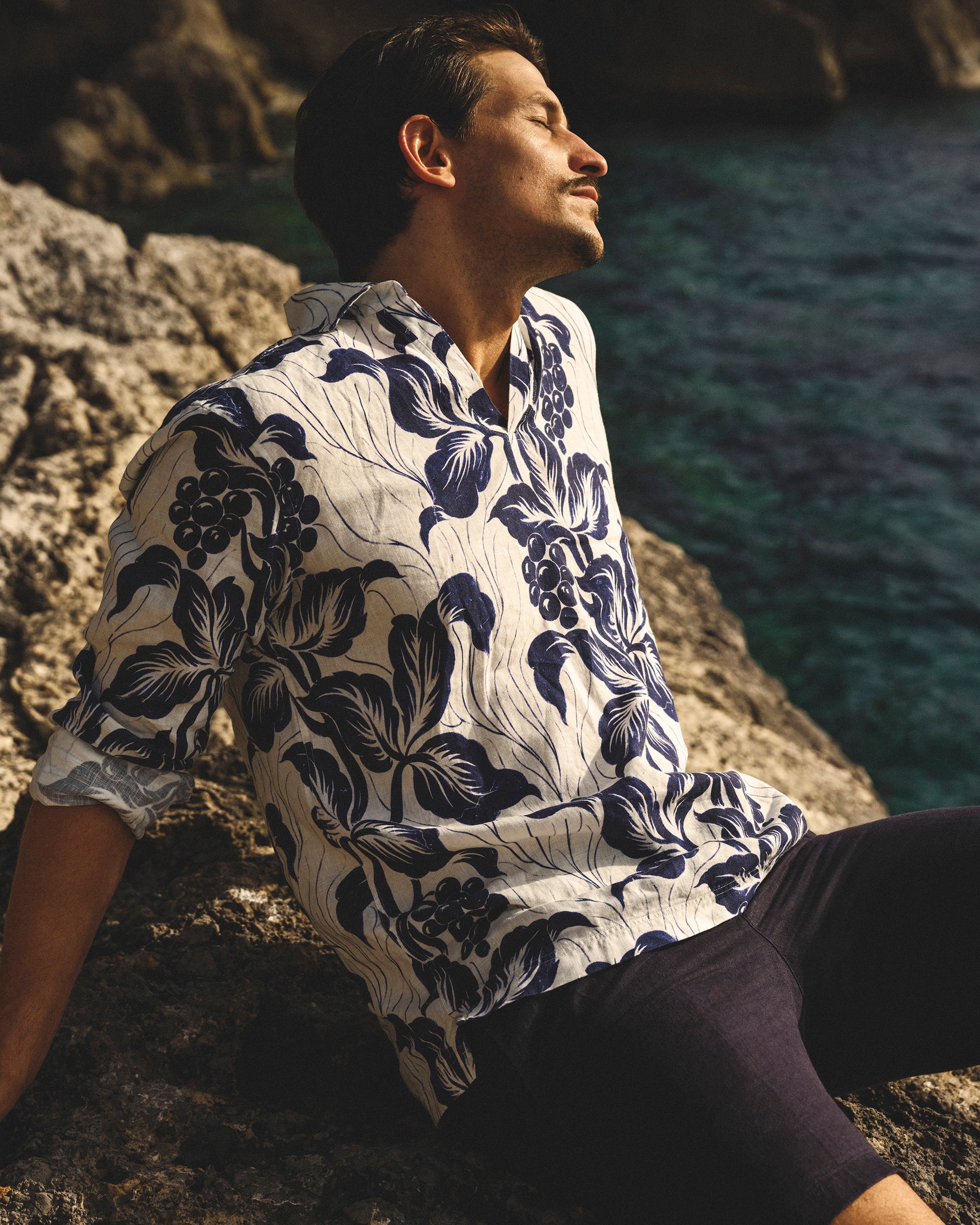Man wearing a floral shirt sitting on a rocky outcrop by the sea.