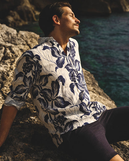 Man wearing a floral shirt sitting on a rocky outcrop by the sea.
