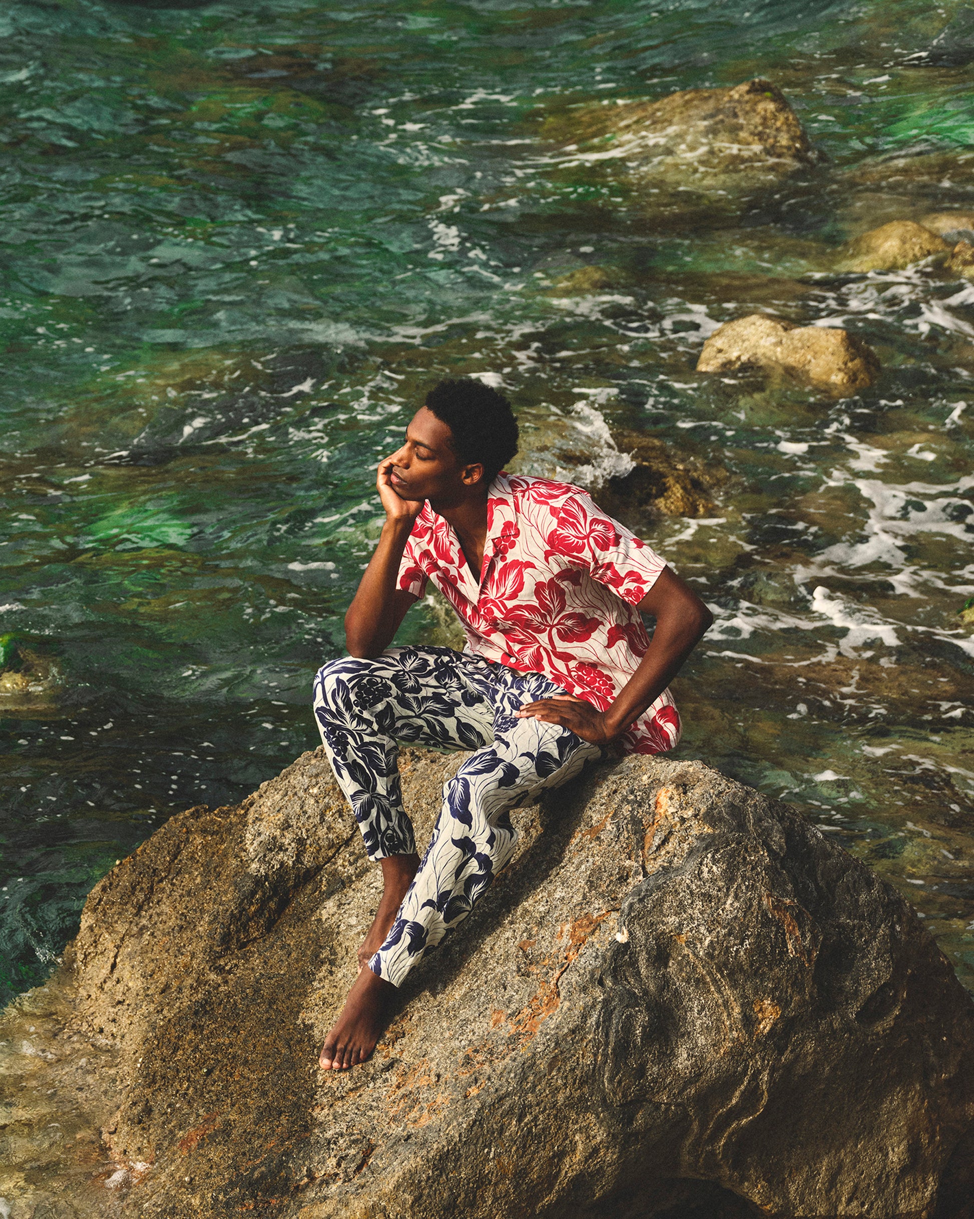 Person sitting on a rock by the water wearing a red floral shirt and patterned pants.