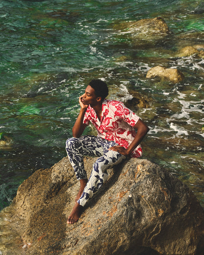 Person sitting on a rock by the water wearing a red floral shirt and patterned pants.