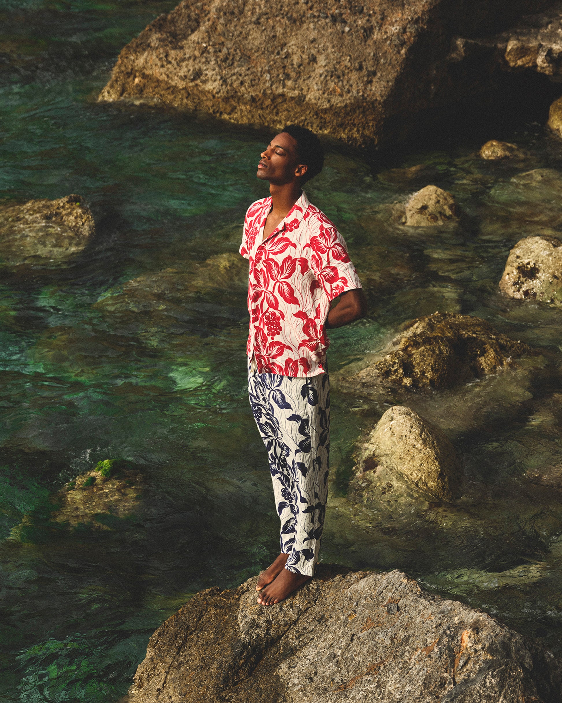 Person standing on a rock by the water wearing a red floral shirt and patterned pants.