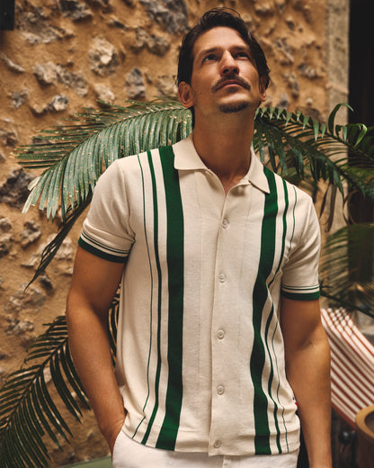 Man wearing a white and green striped shirt with a stone wall and plants in the background