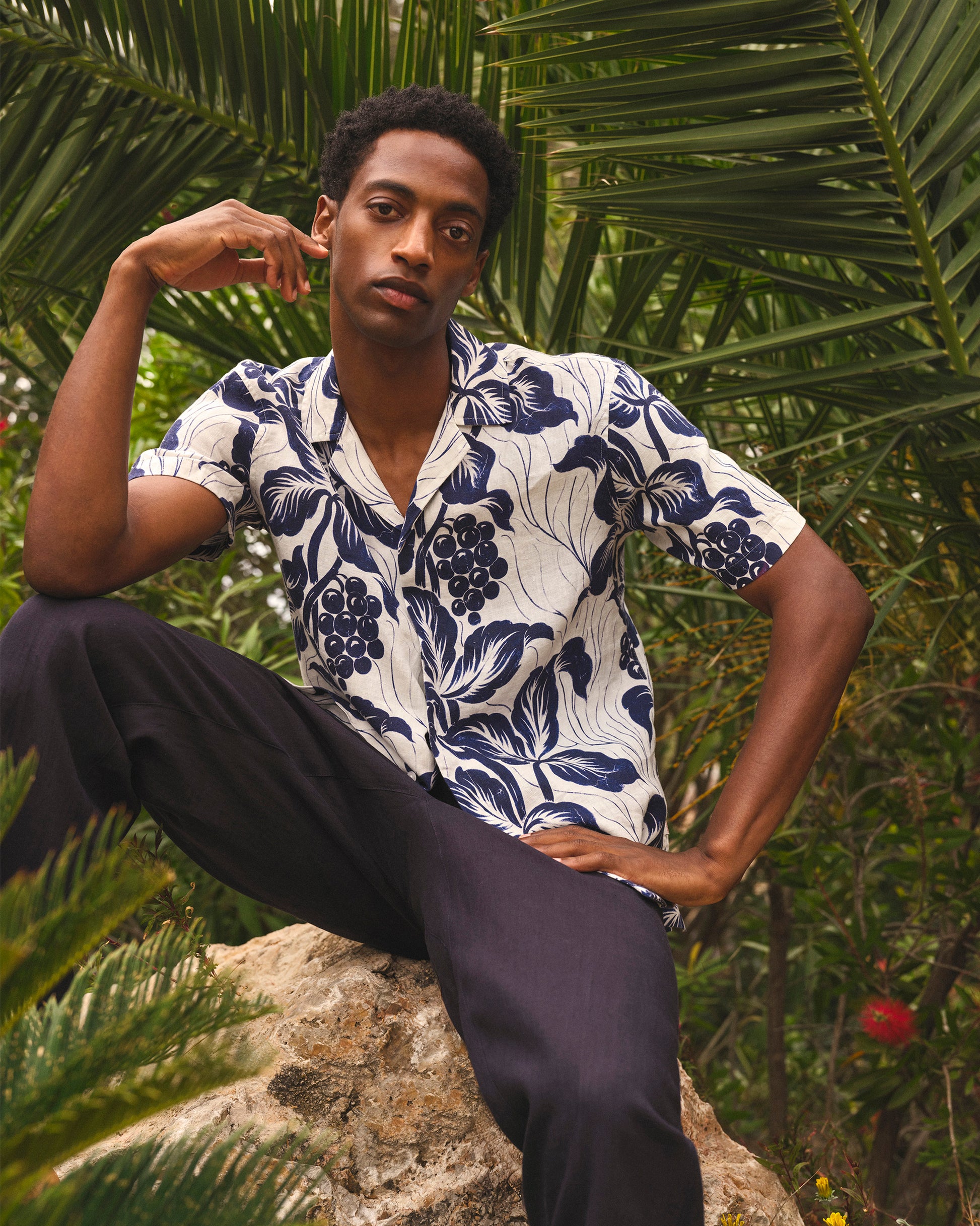 Man wearing a floral shirt sitting on a rock with green foliage in the background