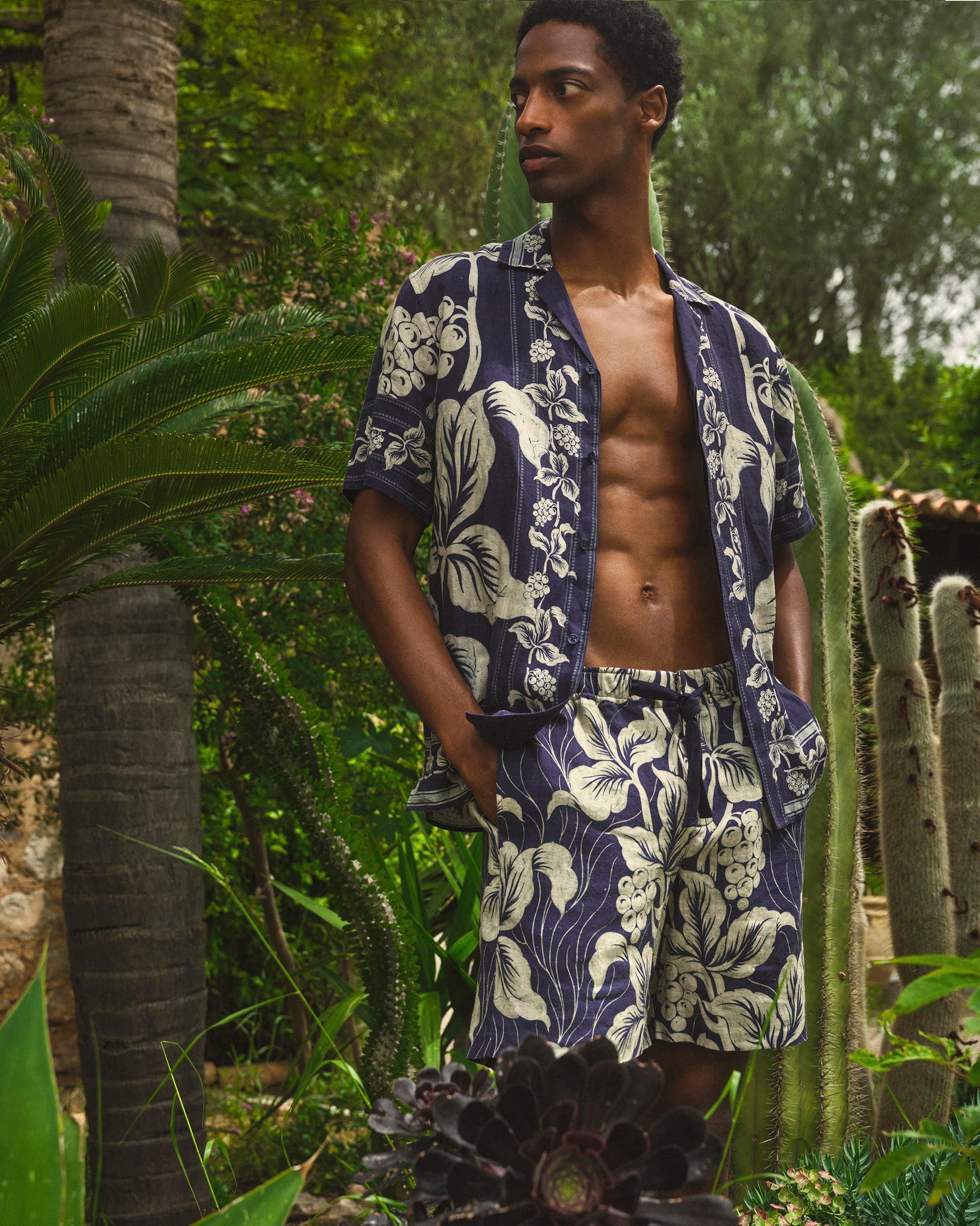 Man wearing a navy blue floral patterned shirt and shorts in a tropical setting with cacti and palm trees.