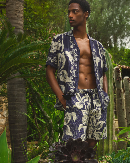 Man wearing a navy blue floral patterned shirt and shorts in a tropical setting with cacti and palm trees.