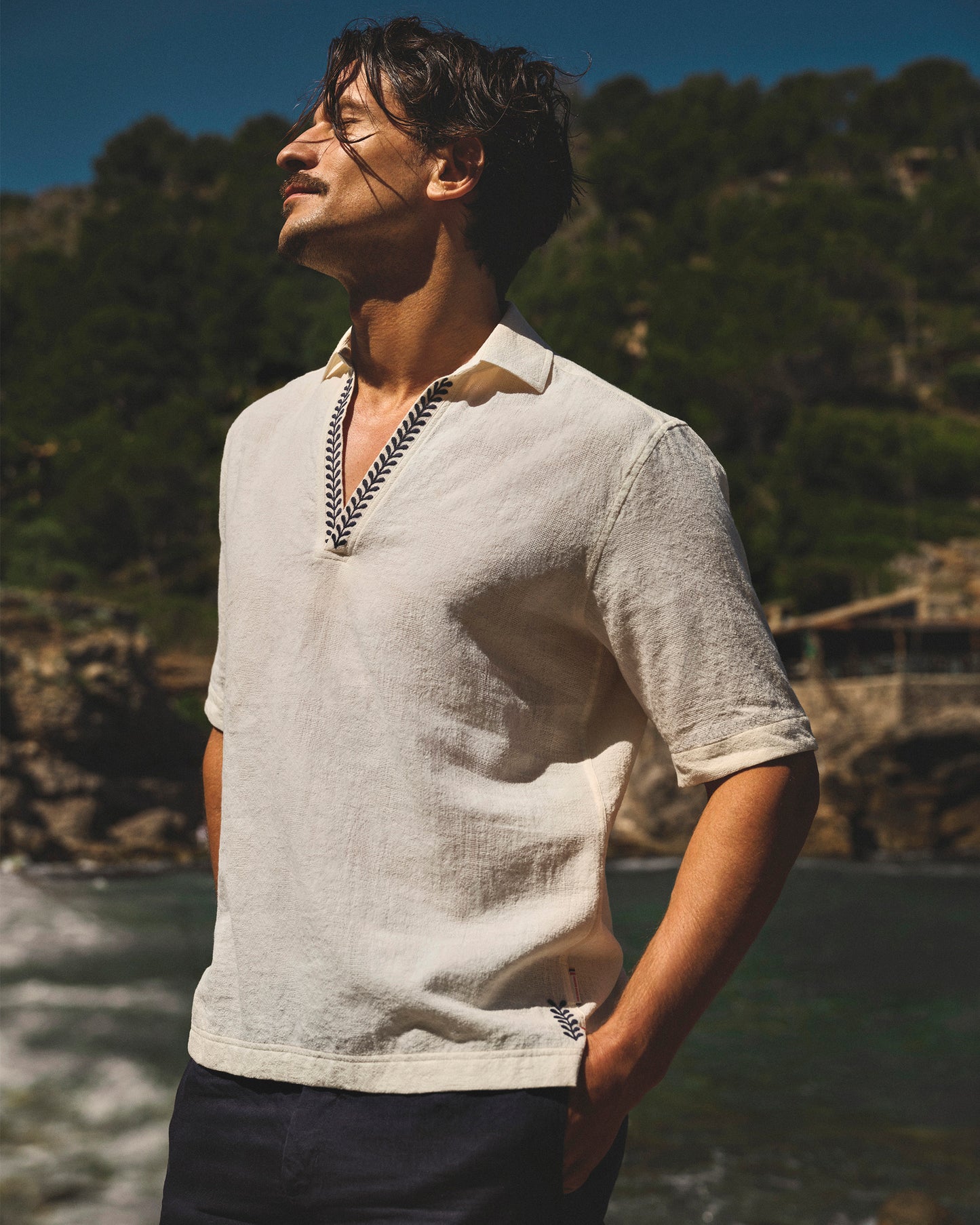 Man wearing a light-colored shirt with a collar, standing by a body of water with trees in the background.