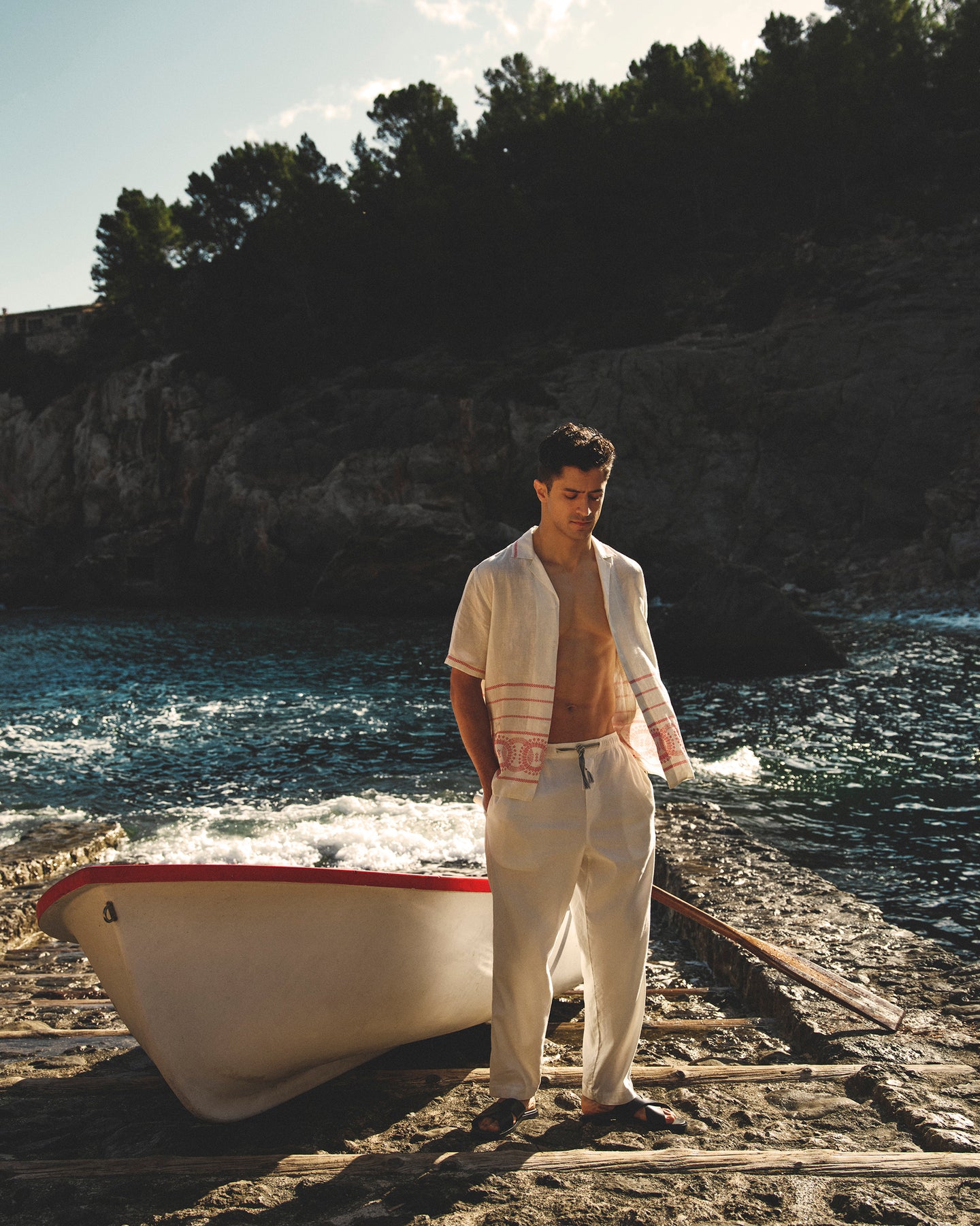 Man standing next to a boat on a rocky shore with water and trees in the background