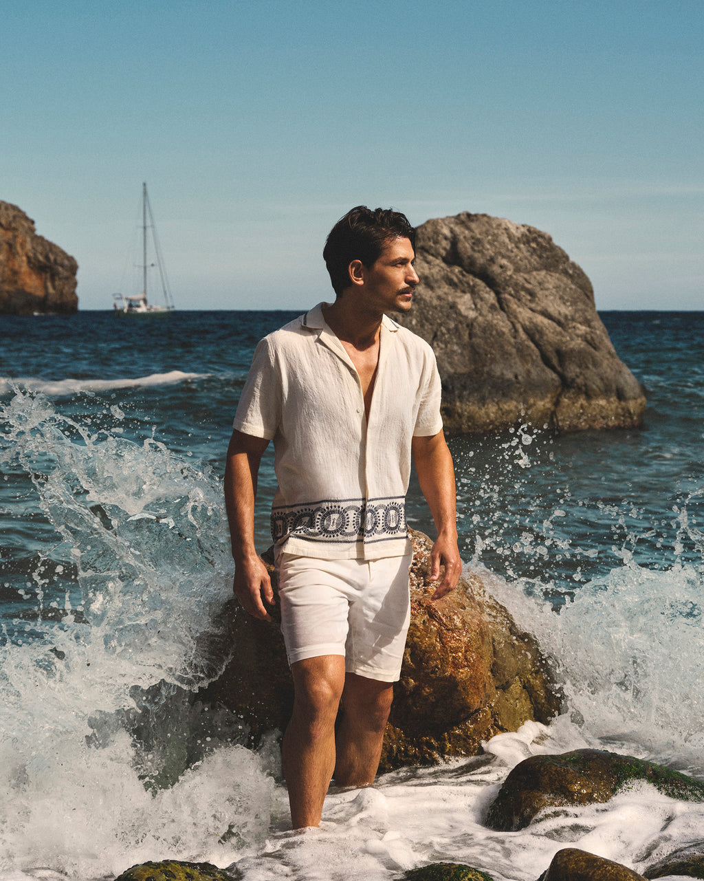Man in white outfit standing on rocks by the sea with a sailboat in the background