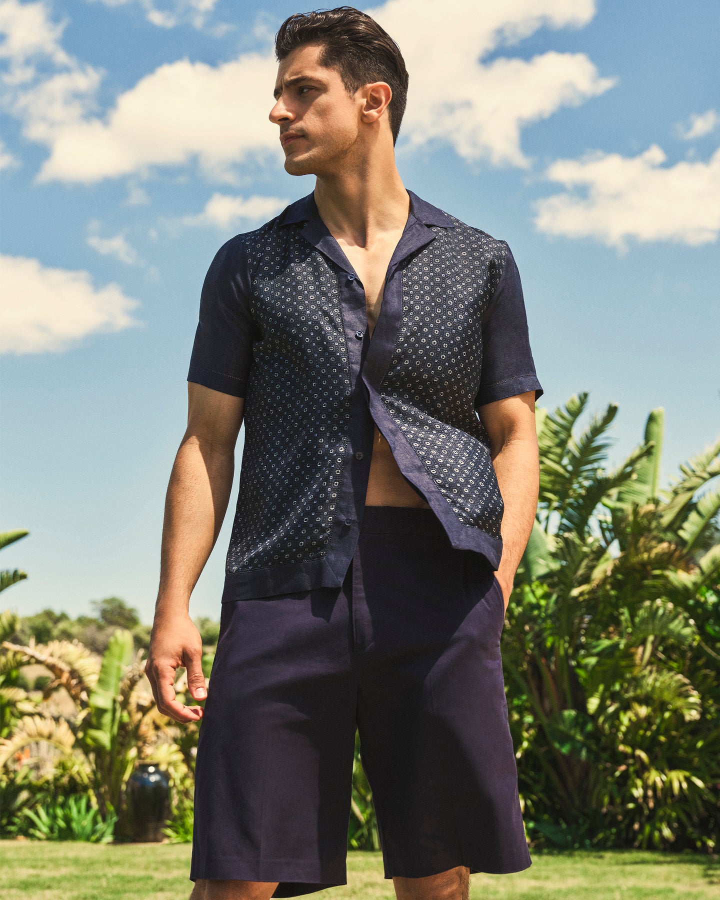 Man wearing a navy patterned shirt and shorts standing outdoors with greenery and blue sky in the background