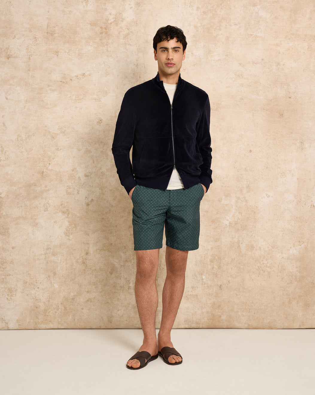 Man wearing a black jacket and green shorts against a beige wall