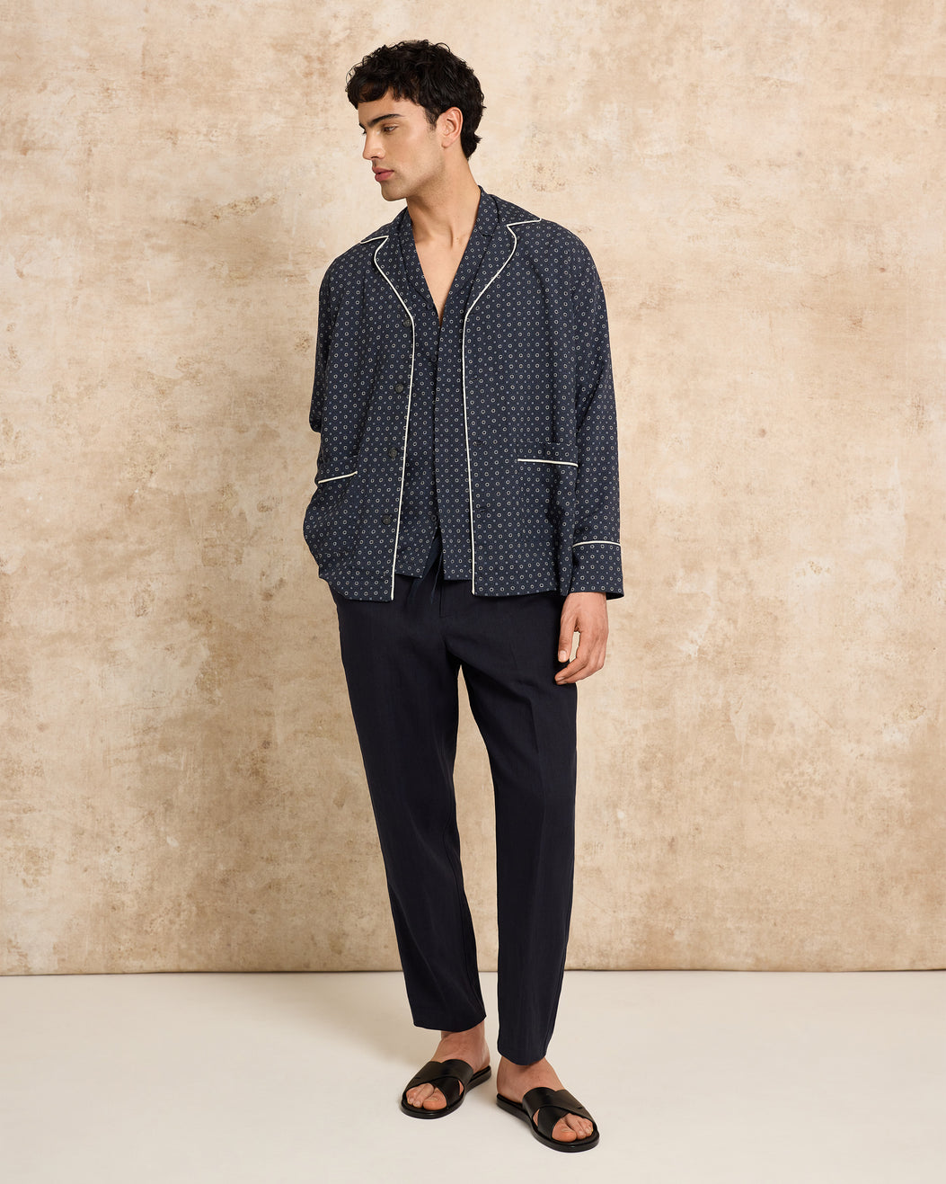 Man wearing a dark patterned pajama set against a beige wall.