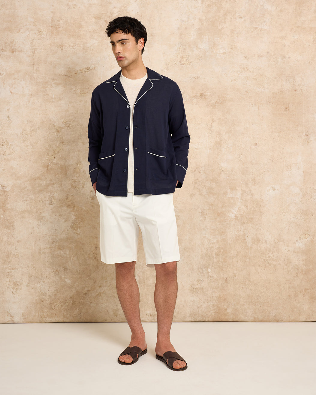 Man wearing a navy jacket and white shorts against a beige wall