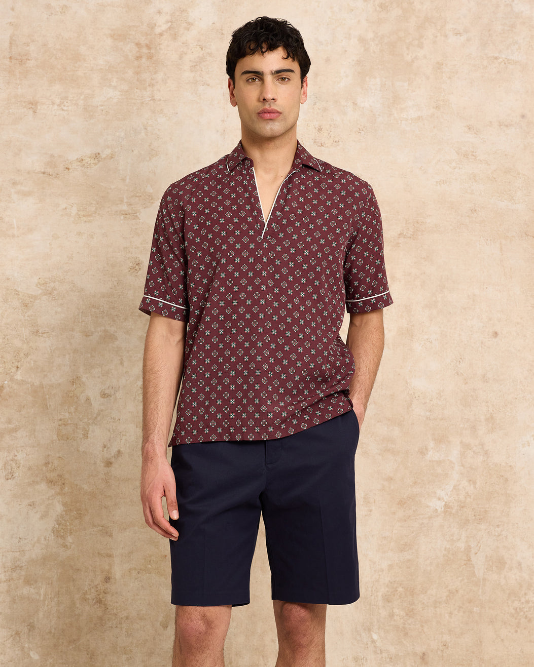 Man wearing a patterned shirt and navy shorts against a beige background