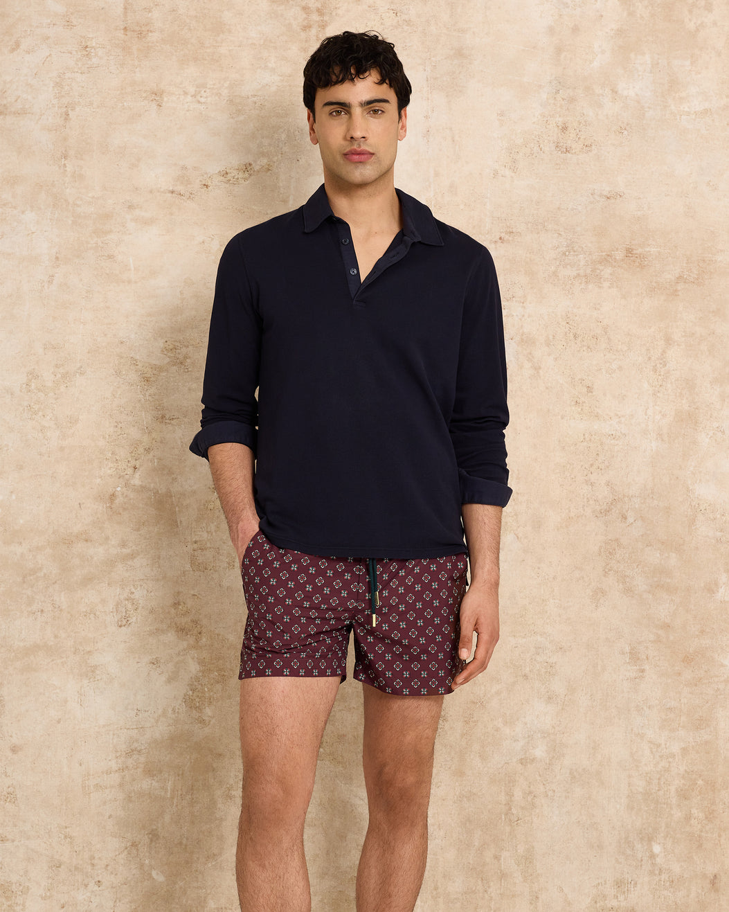 Man wearing a dark long-sleeve shirt and patterned shorts against a beige background
