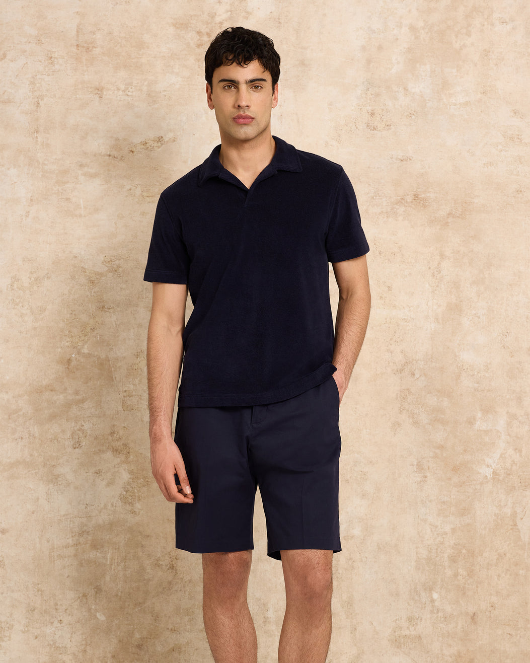 Man wearing a navy polo shirt and shorts against a beige wall