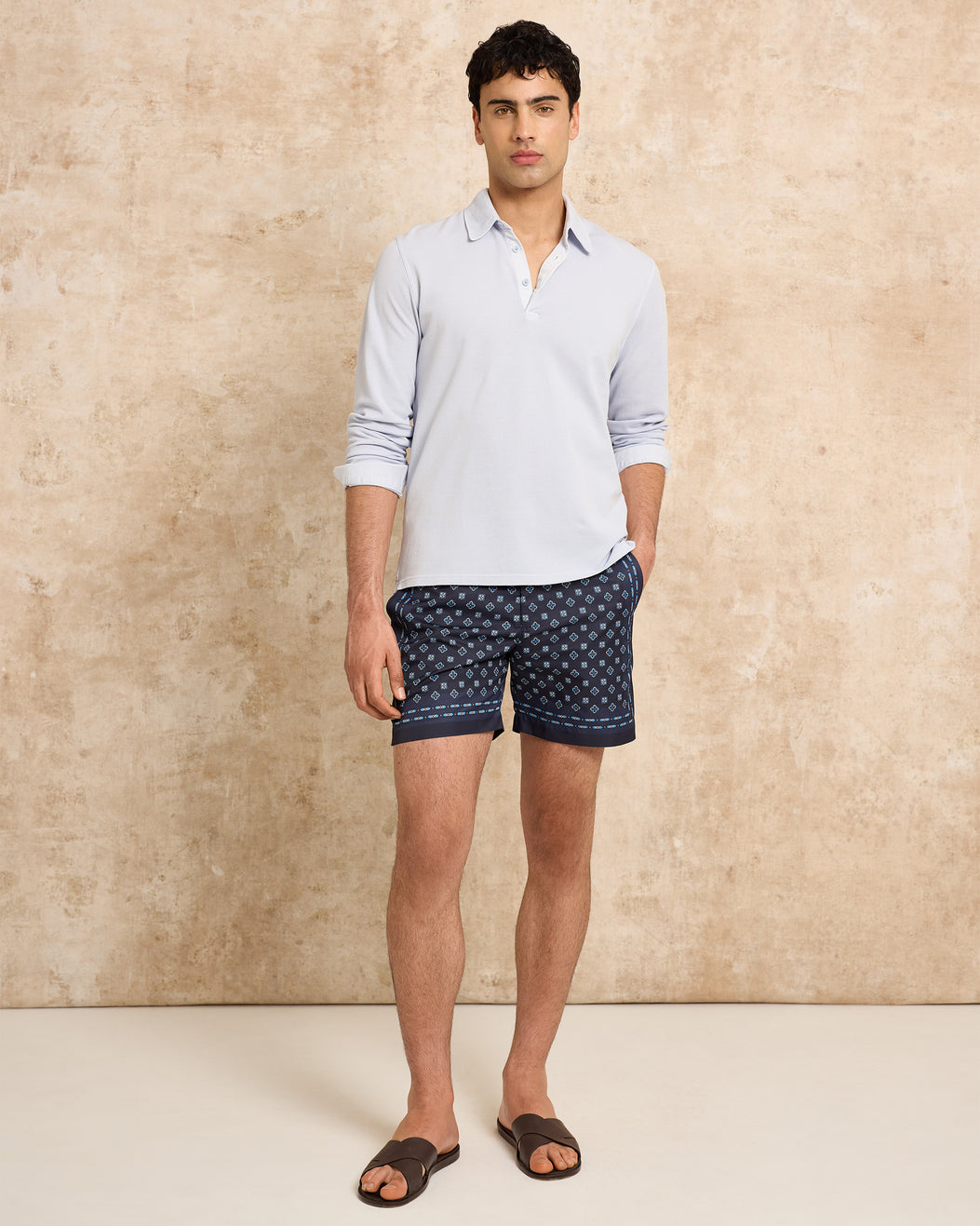Man wearing a light blue polo shirt and navy patterned shorts against a beige background