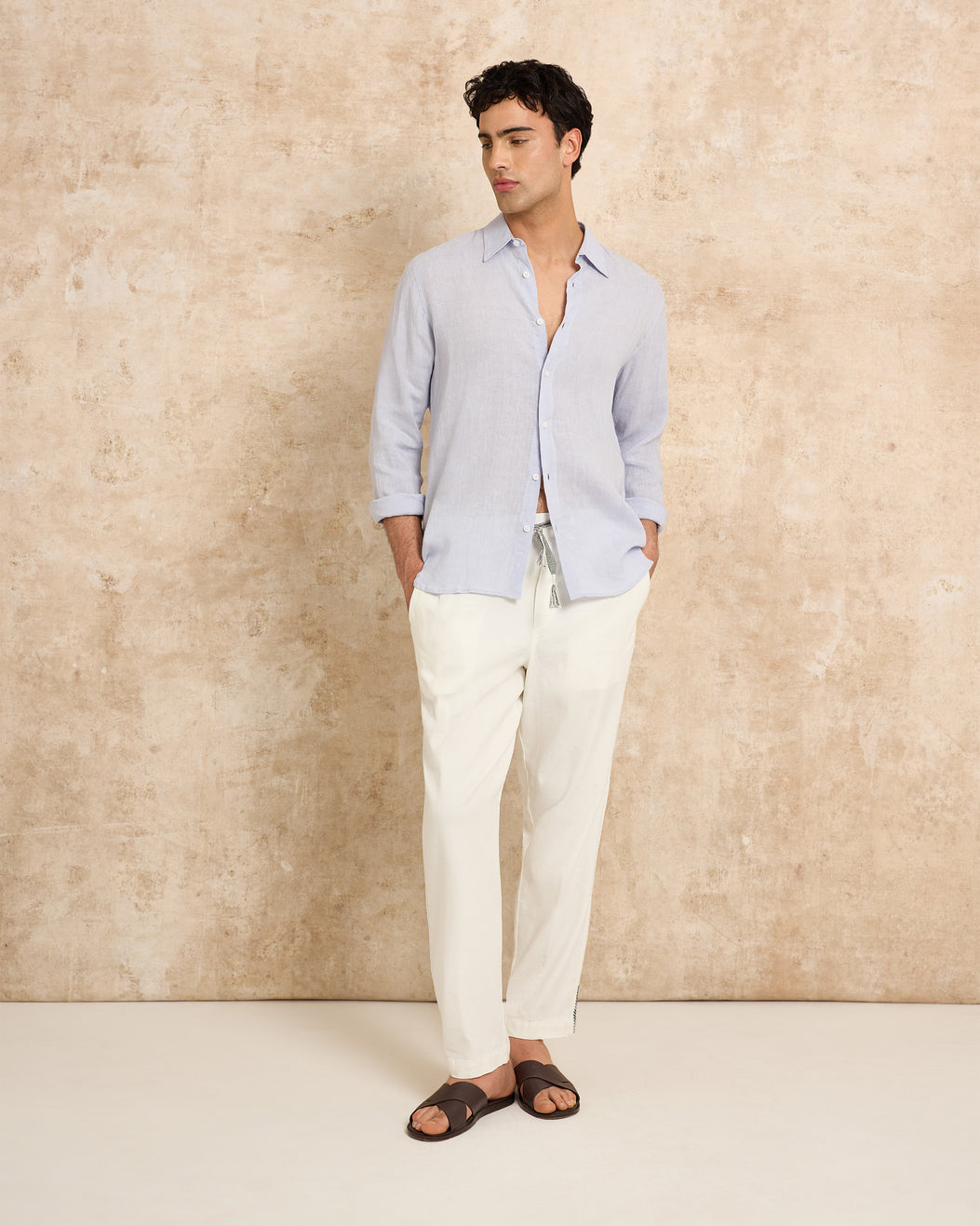 Man wearing a light blue shirt and white pants against a beige wall.