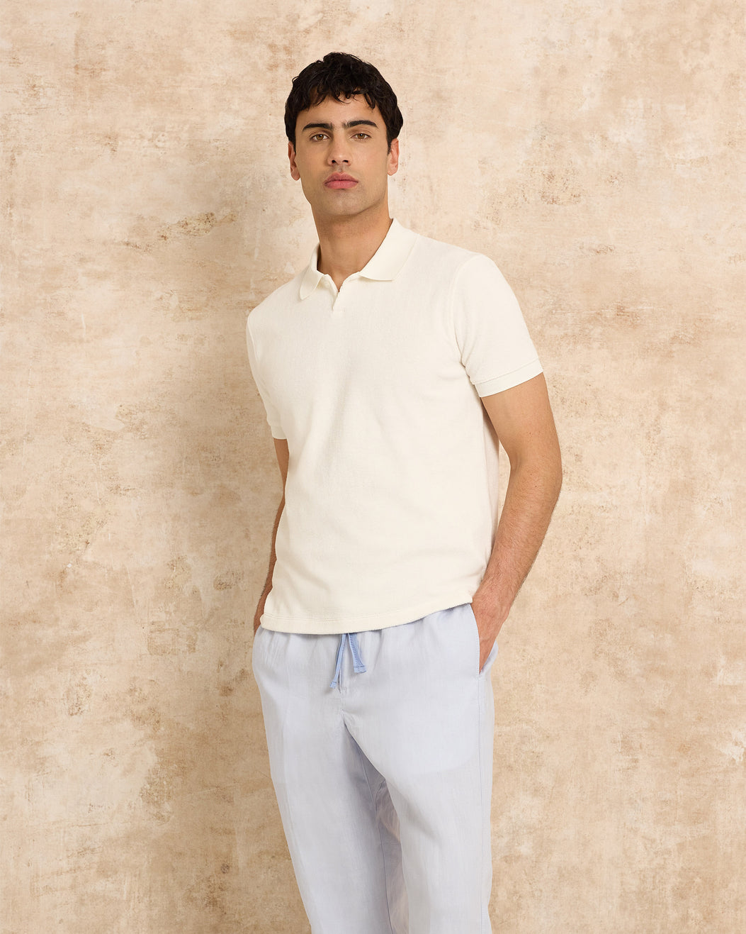 Man wearing a white polo shirt and light blue pants against a beige wall