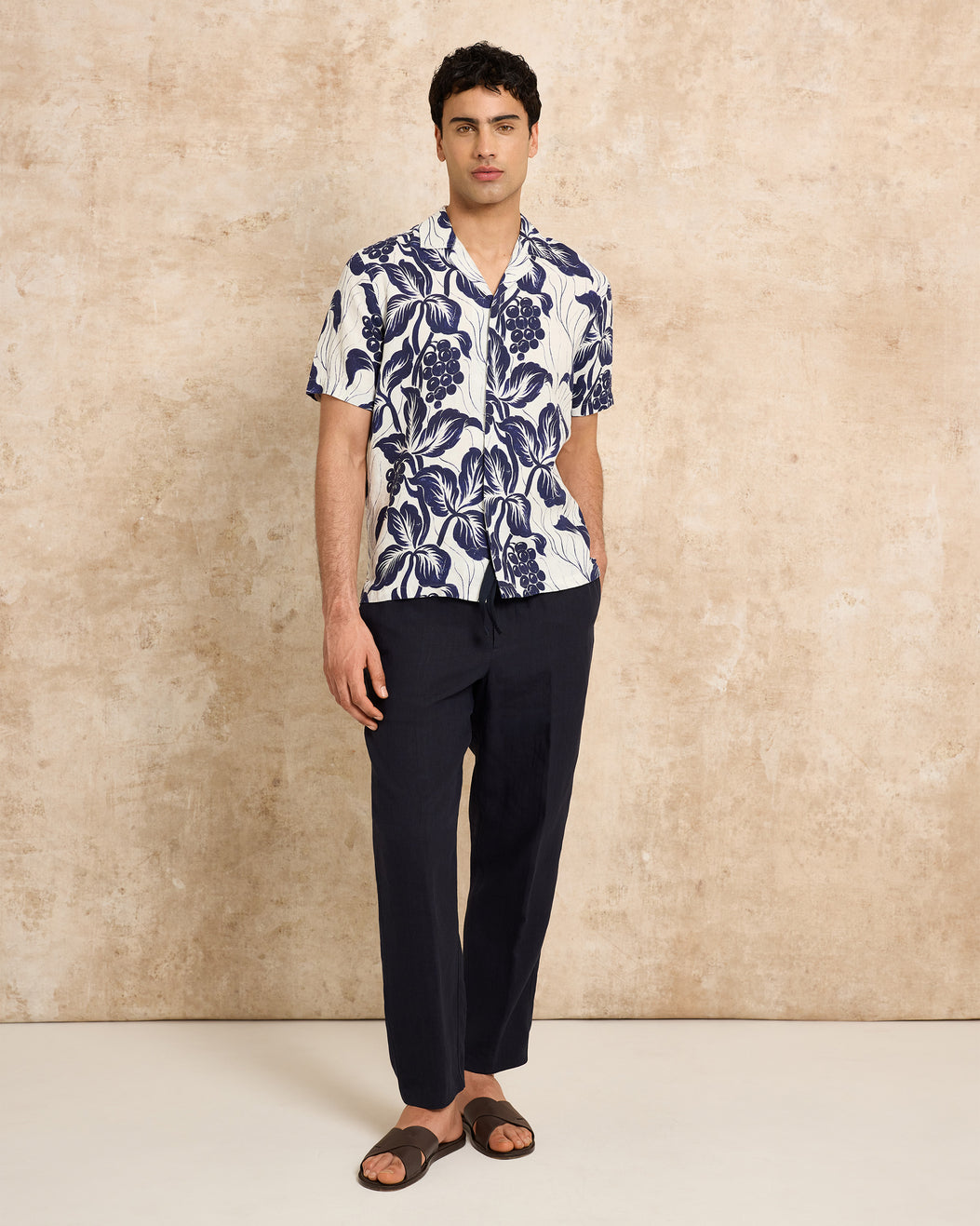 Man wearing a patterned shirt and dark pants against a beige wall