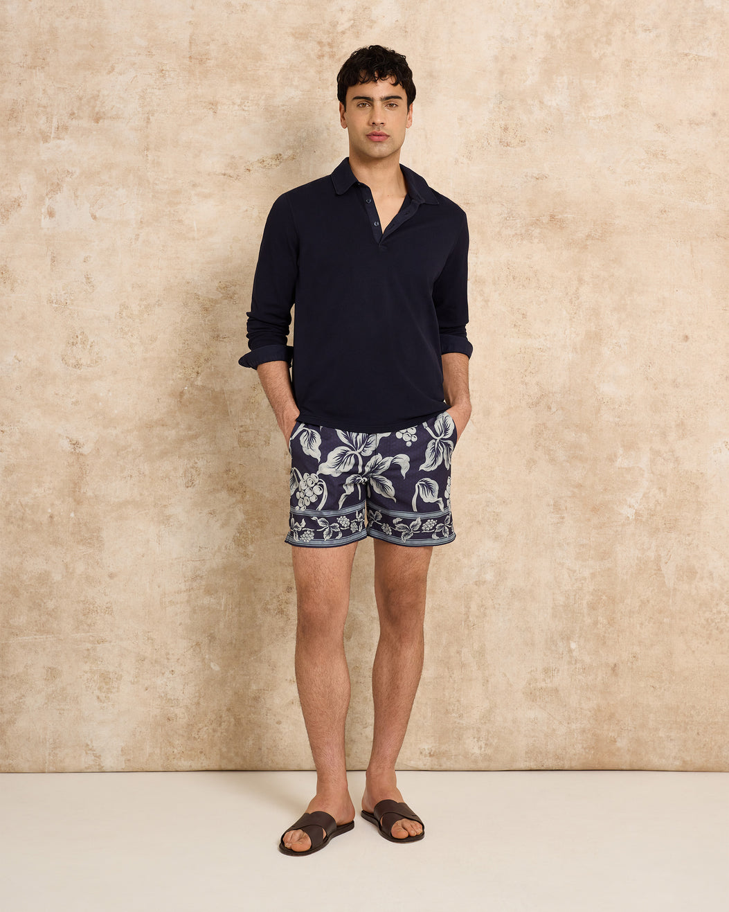 Man wearing a navy shirt and patterned shorts against a beige wall