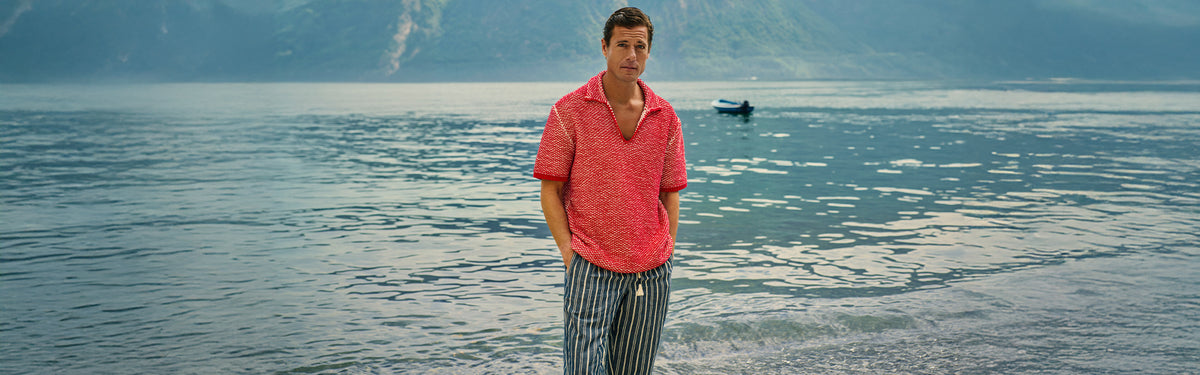 Man wearing red crochet stitch polo shirt with striped trousers against lake background